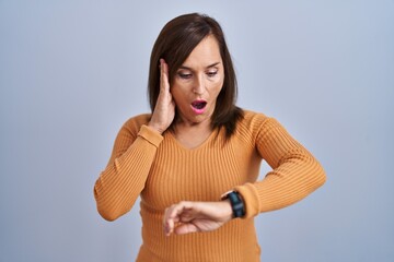 Middle age brunette woman standing wearing orange sweater looking at the watch time worried, afraid...