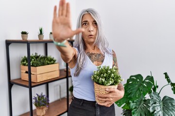 Middle age grey-haired woman holding green plant pot at home doing stop gesture with hands palms, angry and frustration expression