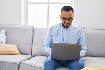 Young hispanic man using laptop and earphones sitting on sofa at home