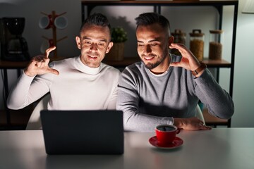 Homosexual couple using computer laptop smiling and confident gesturing with hand doing small size sign with fingers looking and the camera. measure concept.