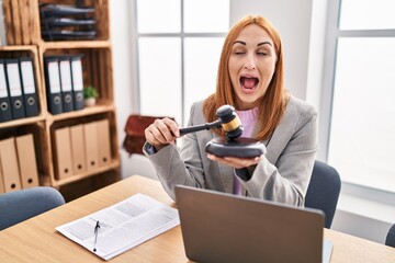Young business woman using gavel angry and mad screaming frustrated and furious, shouting with...