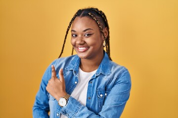 African american woman with braids standing over yellow background cheerful with a smile on face pointing with hand and finger up to the side with happy and natural expression