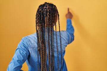 African american woman with braids standing over yellow background posing backwards pointing ahead with finger hand
