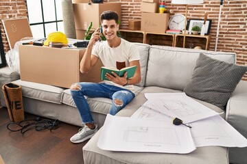 Young hispanic man talking on the smartphone reading book at new home