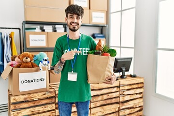 Hispanic man with beard wearing volunteer t shirt at donations point smiling happy pointing with...