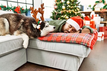 Young hispanic man sleeping lying on sofa with dog by christmas tree at home