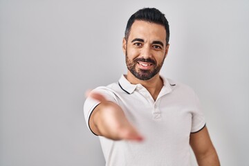 Young hispanic man with beard wearing casual clothes over white background smiling cheerful offering palm hand giving assistance and acceptance.