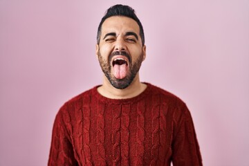 Young hispanic man with beard wearing casual sweater over pink background sticking tongue out happy with funny expression. emotion concept.