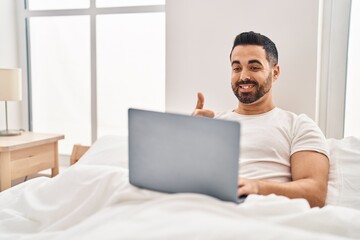 Young hispanic man with beard using computer laptop on the bed smiling happy and positive, thumb up doing excellent and approval sign