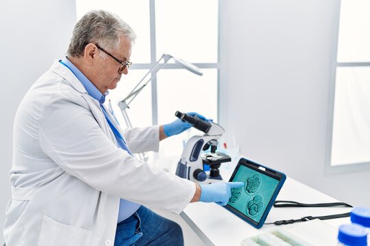 Middle age grey-haired man wearing scientist uniform looking embryology image touchpad at laboratory