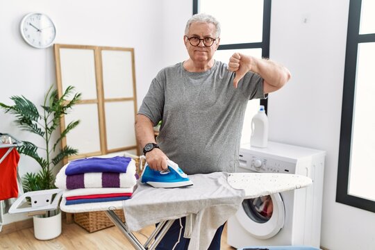 Senior caucasian man ironing clothes at home with angry face, negative sign showing dislike with thumbs down, rejection concept