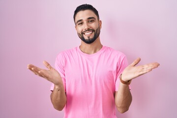 Hispanic young man standing over pink background smiling cheerful offering hands giving assistance and acceptance.