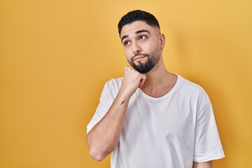 Young handsome man wearing casual t shirt over yellow background with hand on chin thinking about question, pensive expression. smiling with thoughtful face. doubt concept.