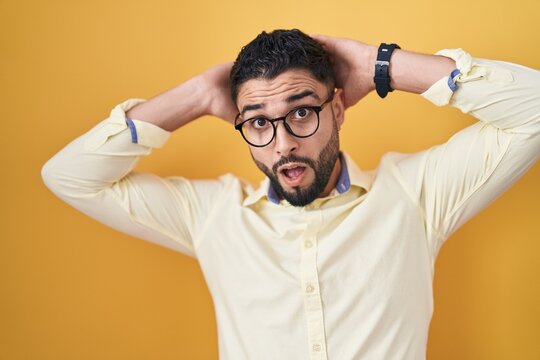 Hispanic young man wearing business clothes and glasses crazy and scared with hands on head, afraid and surprised of shock with open mouth