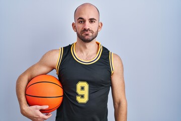 Young bald man with beard wearing basketball uniform holding ball relaxed with serious expression on face. simple and natural looking at the camera.