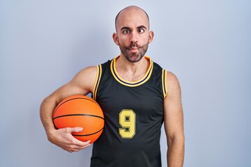 Young bald man with beard wearing basketball uniform holding ball making fish face with lips, crazy and comical gesture. funny expression.