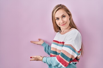 Young blonde woman standing over pink background inviting to enter smiling natural with open hand