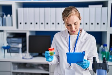 Young blonde woman scientist using touchpad holding urine test tube at laboratory