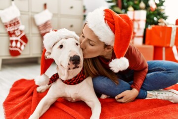 Young caucasian woman kissing and sitting with dog by christmas tree at home