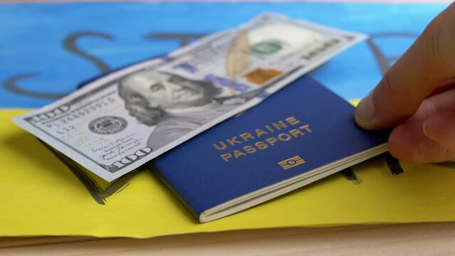 Hand Puts a Biometric Passport of Ukraine with Cash on a Background of Flag. Drawing of yellow, blue Ukrainian flag with title stop the war lie on table. Money, bribe. War, migrants, Russian attack.