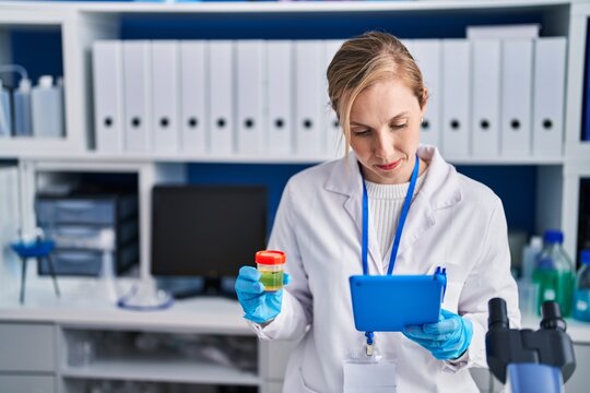 Young blonde woman scientist using touchpad holding urine test tube at laboratory