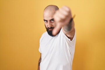 Young hispanic man with beard and tattoos standing over yellow background looking unhappy and angry showing rejection and negative with thumbs down gesture. bad expression.