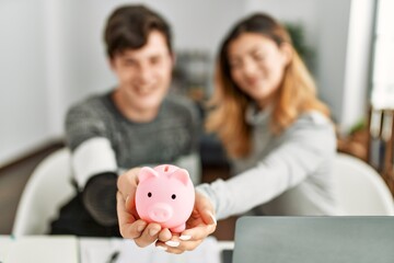 Obraz na płótnie Canvas Young caucasian couple smiling happy holding piggy bank sitting on the table at home.