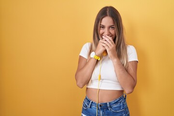 Young blonde woman standing over yellow background wearing headphones laughing and embarrassed...
