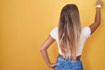 Young blonde woman standing over yellow background wearing headphones posing backwards pointing ahead with finger hand