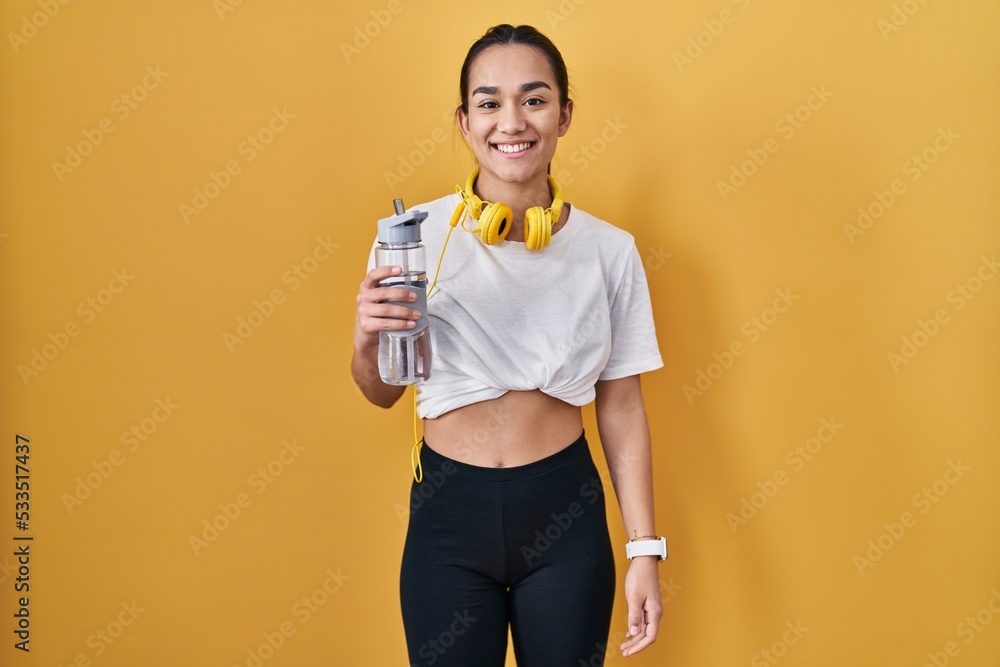 Poster young south asian woman wearing sportswear drinking water with a happy and cool smile on face. lucky