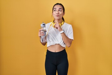 Young south asian woman wearing sportswear drinking water thinking concentrated about doubt with...