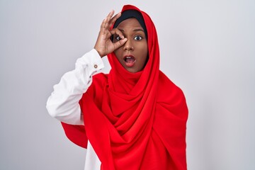 Young arab woman wearing traditional islamic hijab scarf doing ok gesture shocked with surprised face, eye looking through fingers. unbelieving expression.