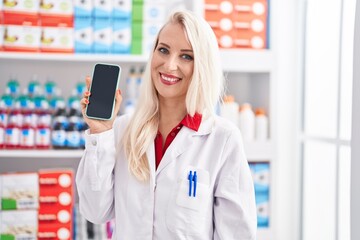 Caucasian woman working at pharmacy drugstore showing smartphone screen looking positive and happy standing and smiling with a confident smile showing teeth