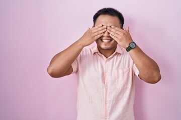 Chinese young man standing over pink background covering eyes with hands smiling cheerful and funny. blind concept.