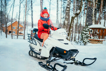 portrait of a girl in a red hat and scarf sits on a snowmobile in a snowy forest on a cold day