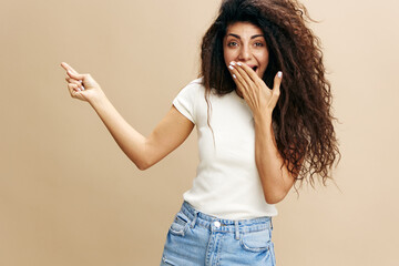 SEASONAL SALE OFFER. Excited beautiful Latin female in white t-shirt with afro say Look at this, close mouth, show copy space, free place for design ad. Fashion. Studio shoot over beige background