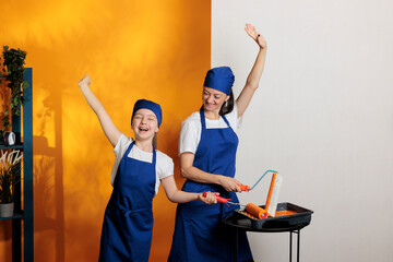 Cheerful people painting walls with orange color paint and roller brush, having fun with house renovation. Mother and little child using paintbrush and redecoration tools to do housework improvement.