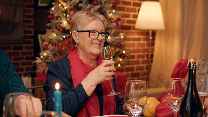 Festive diverse people clinking glasses while enjoying Christmas dinner table at home. Happy family members sitting in living room and clinking glasses while celebrating winter holiday together.