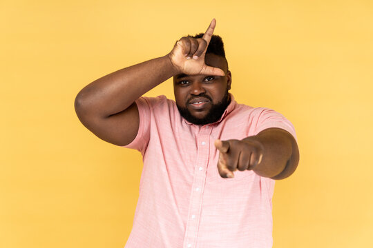 Portrait of handsome sad frustrated man wearing pink shirt showing looser gesture and pointing at camera, looking with grumpy face. Indoor studio shot isolated on yellow background.