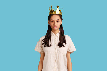 Arrogant selfish woman with black dreadlocks egoistically looking at camera, posing with crown on head, pretending to be queen, wearing white shirt. Indoor studio shot isolated on blue background.