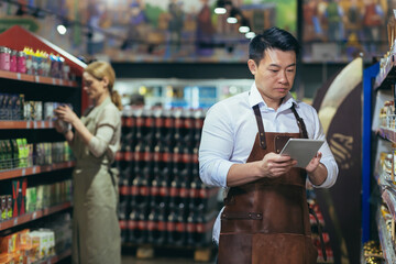 Portrait of young handsome Asian man, shop owner, supermarket worker. Standing in an apron near the...