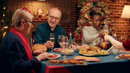 Happy elderly man talking with wife while enjoying Christmas dinner table with close family members. Multiethnic festive people celebrating traditional winter holiday while eating home cooked food.