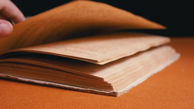 Female Fingers Leafing Through Pages of Old Shabby Book on a Black Background. Reading. Rough papyrus paper on a thick antique book from yesteryear. Orange color. Education. Knowledge. Mystic.