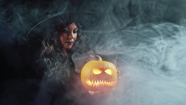 Mysterious black witch with steaming pumpkin as head of jack-o-lantern on dark backdrop. Scary symbol of Halloween, masquerade costume, party decoration. Magic, classic hag