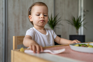 One baby girl small caucasian toddler eating while sit at the table alone at home copy space little child having lunch healthy meal in room in day taking fork