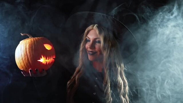 Mysterious black witch with steaming pumpkin as head of jack-o-lantern on dark backdrop. Scary symbol of Halloween, masquerade costume, party decoration. Magic, classic hag