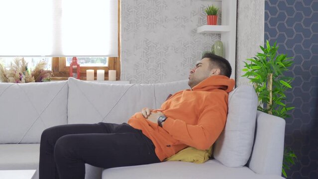 Young man with stomach ache at home.
Young man walks around the house holding his stomach and sits on the sofa and feels pain.
