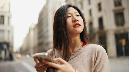 Asian woman looking pensive while exploring new city alone. Female Asian tourist walking along beautiful street with smartphone in hands
