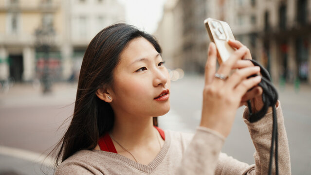 Portrait of pretty Asian woman taking photos of old architecture using smartphone for it on the street. Female Asian tourist exploring European city