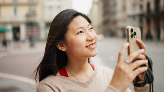 Portrait of pretty long haired Asian woman taking photos of old architecture looking happy while using smartphone on the street